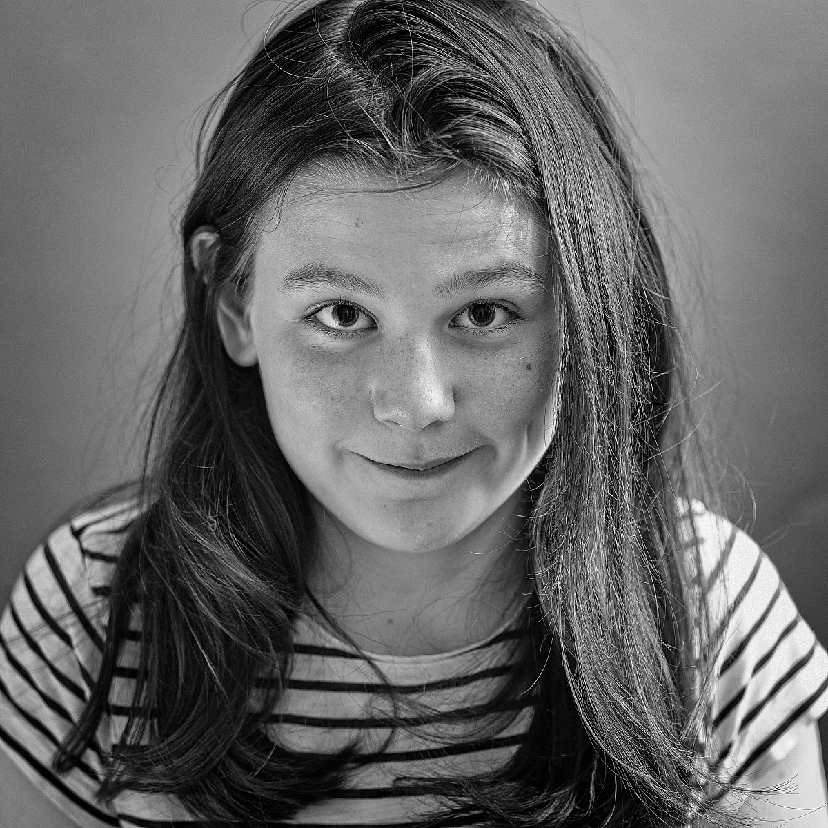 A black-and-white portrait of a person with long hair and a slight smile. They are wearing a striped shirt and looking directly at the camera. The background is plain and out of focus.