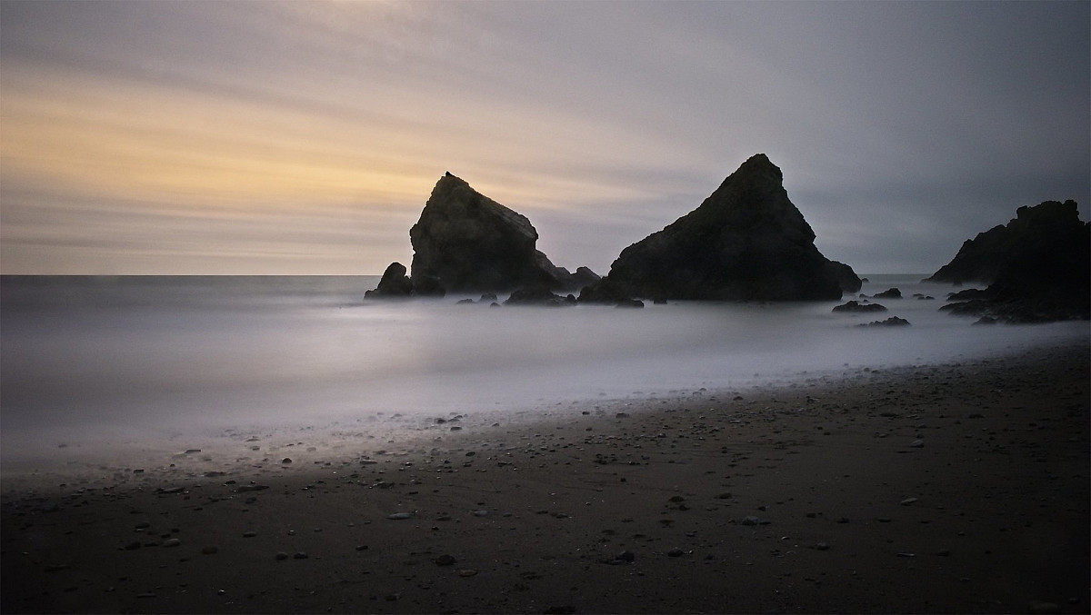 A serene beach scene at dusk featuring large, dark rock formations jutting out from calm water. The sky is painted with soft hues of purple, orange, and grey, creating a tranquil atmosphere. The shore is covered with small pebbles and sand.
