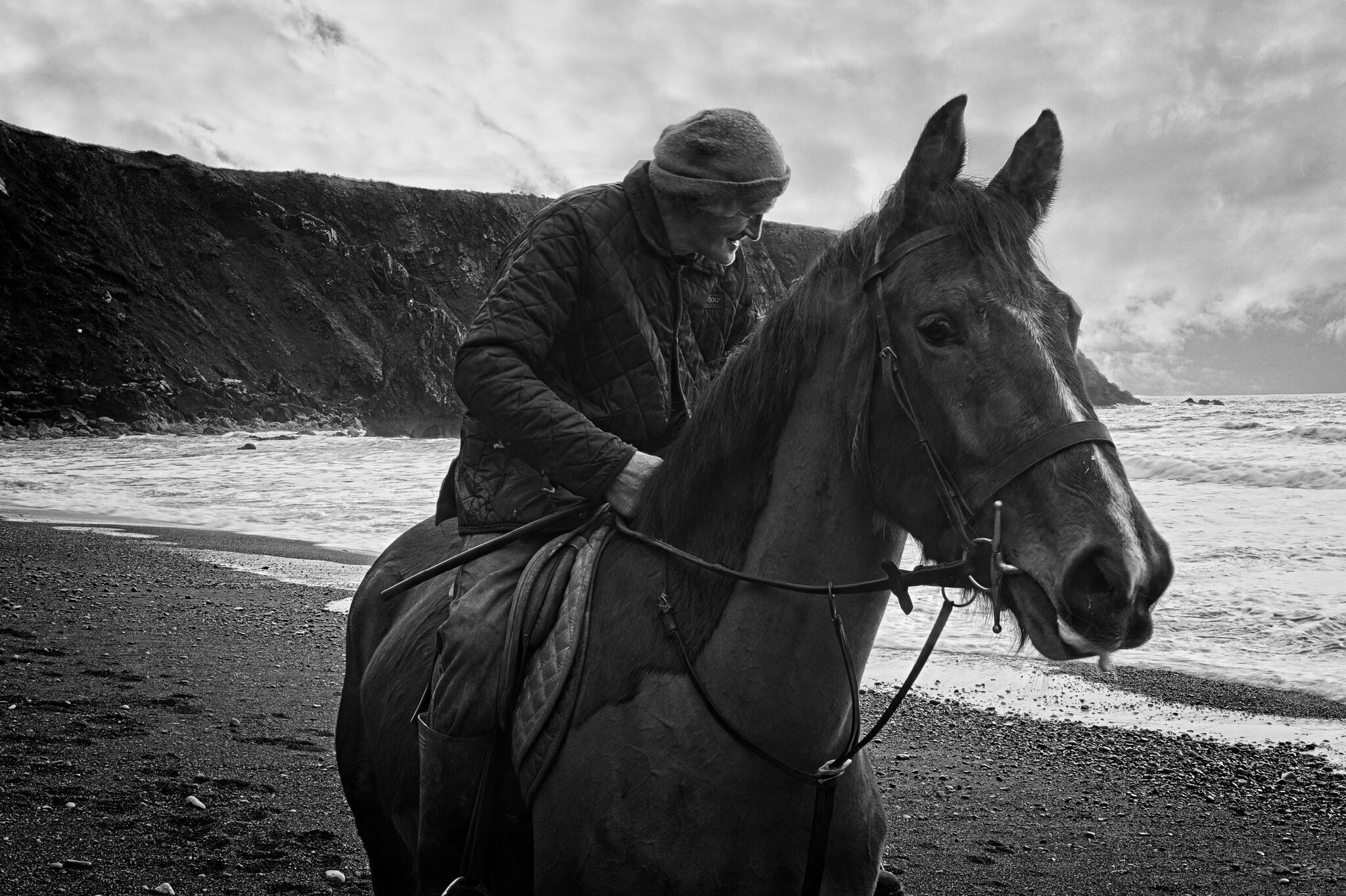 A person wearing a beanie and quilted jacket is sitting on a horse on a rocky beach. The ocean waves are visible in the background, and a cliffside extends along the shoreline. The image is in black and white.