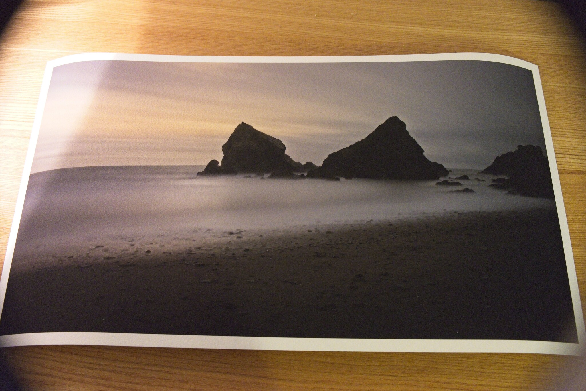 A printed photograph lies on a wooden surface. The image depicts a serene beach scene with dark, jagged rocks jutting out of calm ocean water under a cloudy sky. The photo features a long exposure effect, creating a smooth, misty appearance around the rocks.