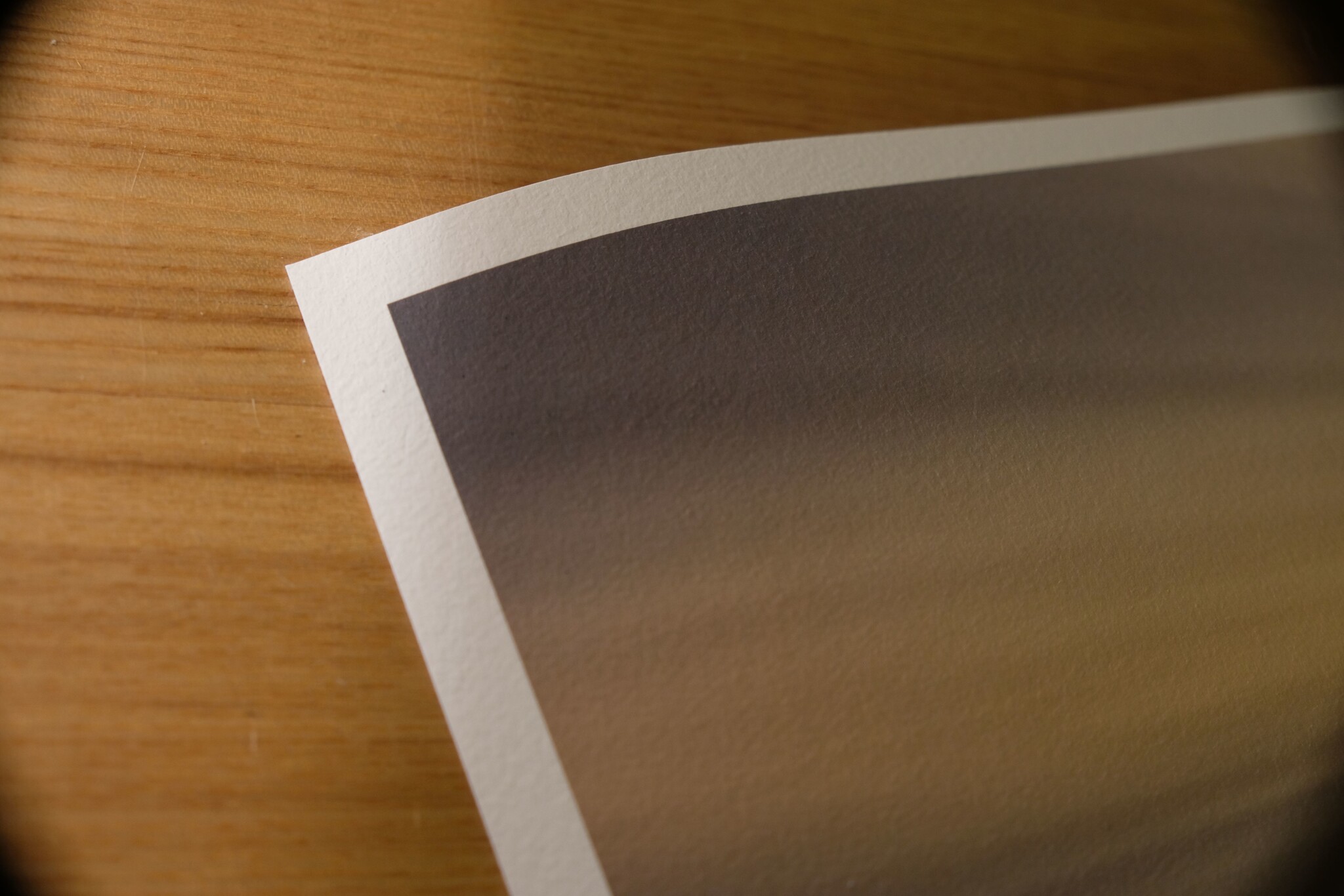 A close-up of the corner of a printed photograph or document lying on a wooden surface. The image appears to have a gradient of warm colors and the paper has a slight curve at the edge.