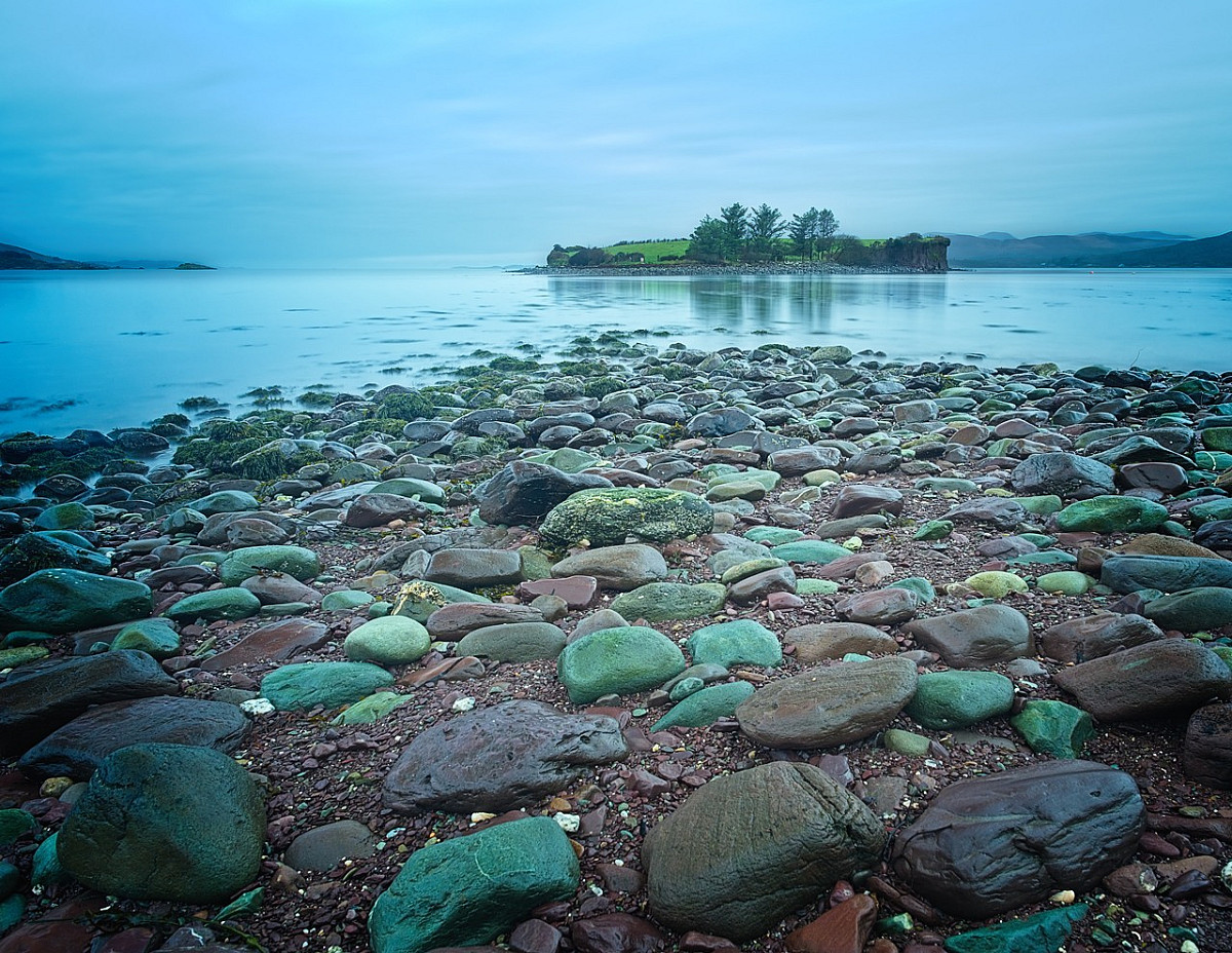Rocky shoreline leading to a calm sea with a small island in the distance. The island is covered with green grass and a few trees. The sky is overcast, casting a bluish tone over the scene.