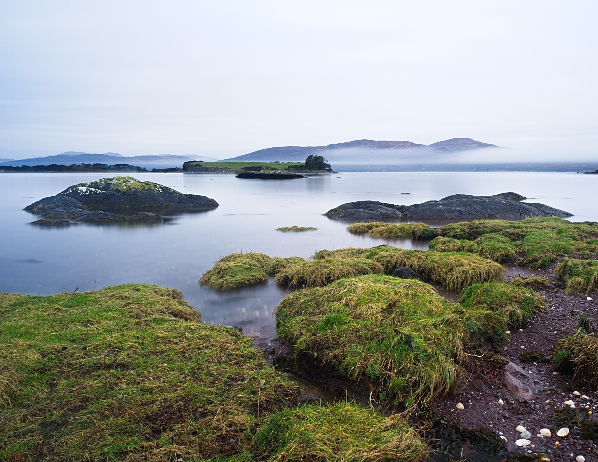 A calm body of water with small grassy islands and rocky outcrops. The scene is tranquil under a grey, overcast sky. In the distance, there are mist-covered mountains, adding a sense of depth and serenity to the landscape.