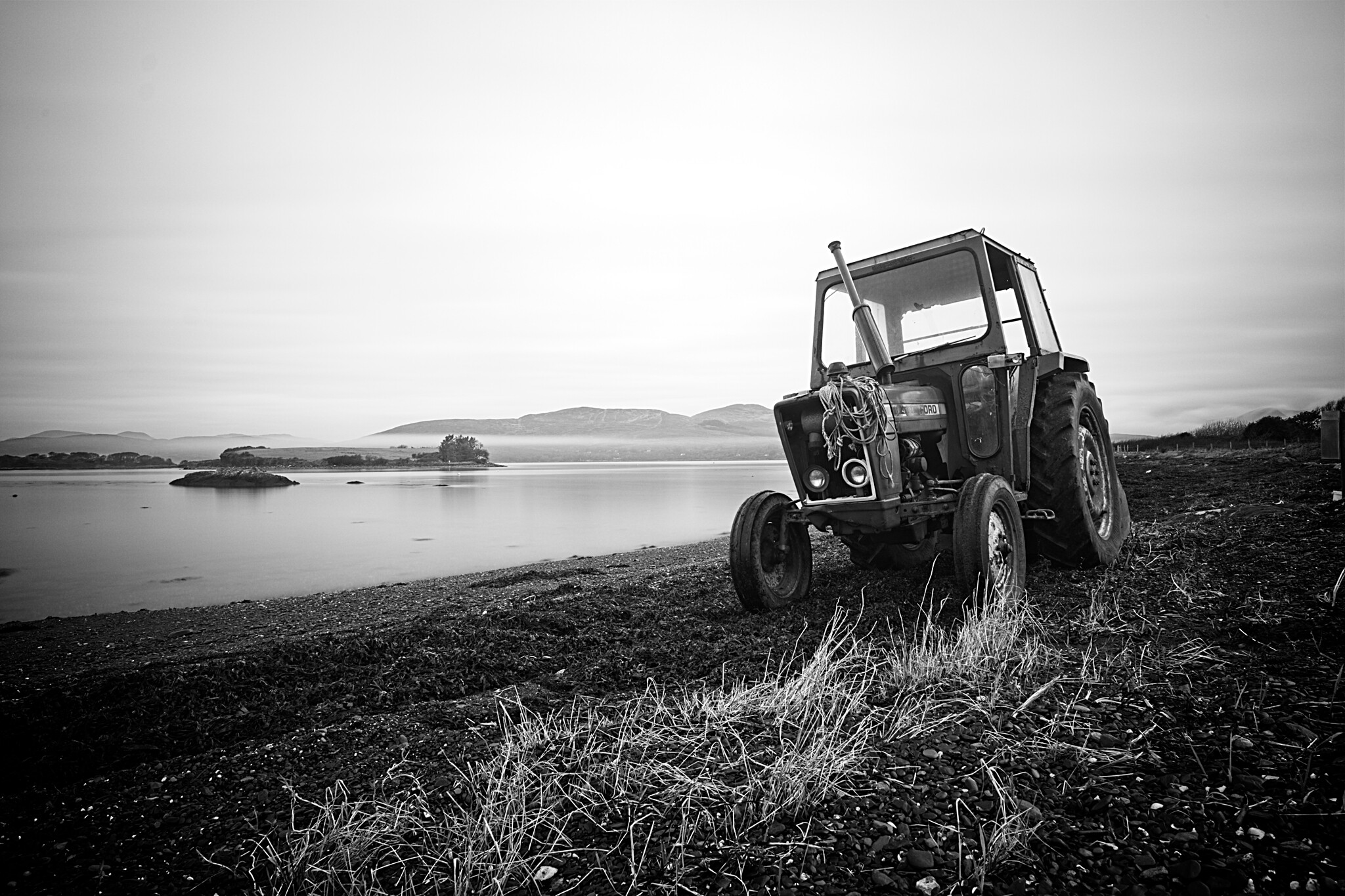 Black and white photo of a tractor parked on a grassy shore with a calm body of water and mountainous terrain in the background. The sky is overcast, and the tractor appears weathered. Small islands are visible in the distance.
