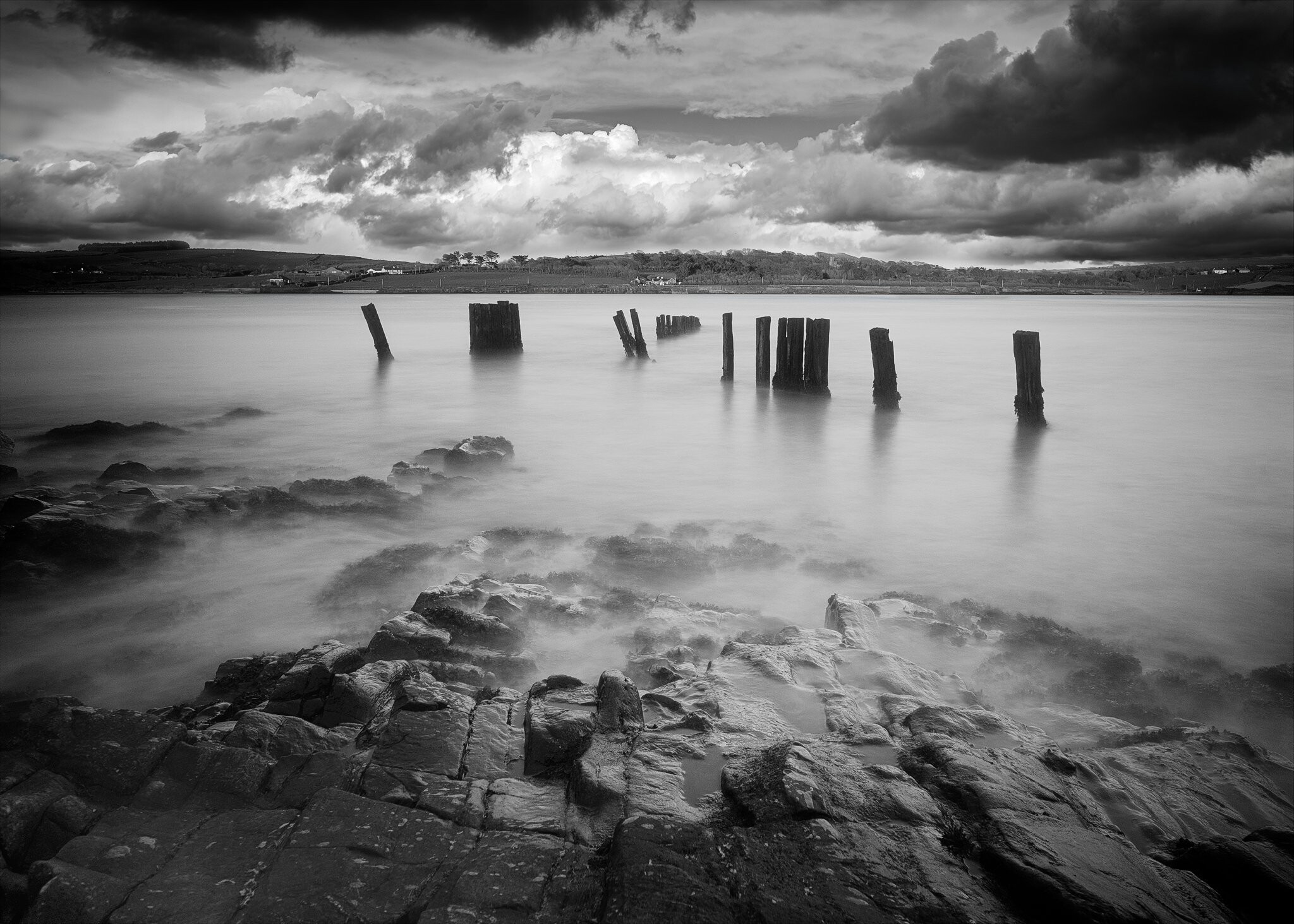 Black and white photo of a rocky shoreline with calm water. Wooden posts, remnants of an old pier, stand in the water. Cloudy sky and distant landscape of rolling hills are visible in the background. The overall atmosphere is serene and slightly moody.